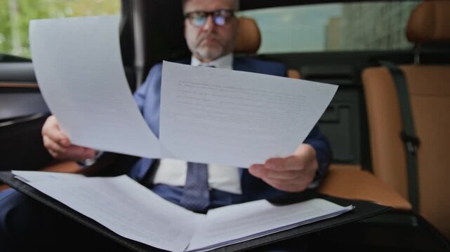 Middle aged Caucasian businessman sitting in back seat of luxury car and reviewing financial documents. Clip is ideal for corporate finance, executive travel, and wealth management marketing