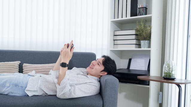 Young Asian man relaxing on sofa browsing smartphone during remote work break in modern home office