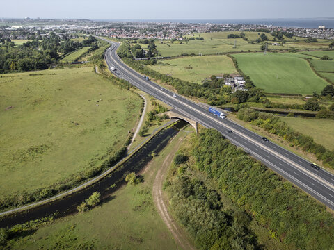Aerial view of a highway crossing over a canal surrounded by green fields and trees near the city in Lancaster, England, United Kingdom.