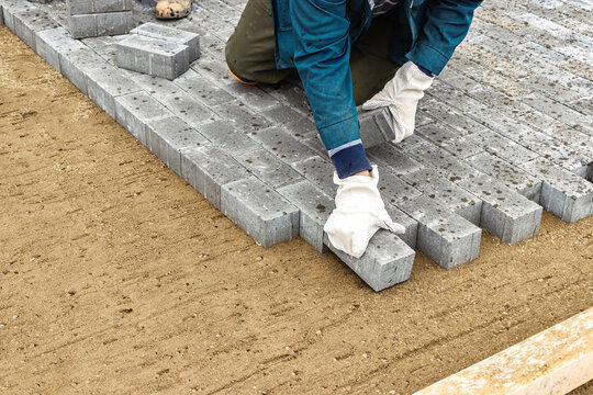 Construction worker laying gray bricks on a sandy foundation in an outdoor setting during daylight hours