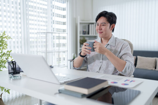 Young Asian businessman holding coffee mug during video conference call at modern home office workspace with copy space