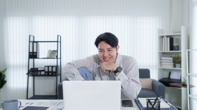 Young Asian businessman smiling while working on laptop in modern home office during hybrid work day
