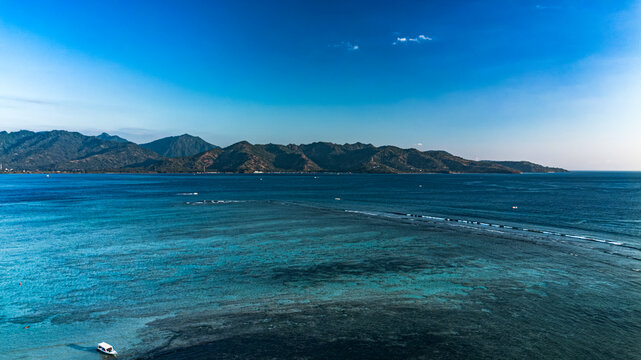 Aerial view of Gili Trawangan surrounded by turquoise waters, coral reefs, and distant mountains under a clear blue sky in Pemenang, West Nusa Tenggara, Indonesia.