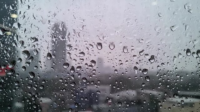 Raindrops covering a car window with a blurred urban scene beyond, creating a moody atmospheric background that evokes rainy weather, travel, and urban life.