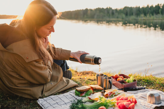 Woman pouring a hot drink from a thermos during an outdoor picnic by a peaceful lake, enjoying fresh food, warm sunset light and relaxing time in nature