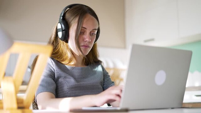 Woman typing on laptop wearing headphones. Remote worker uses computer at home desk. Freelancer works on laptop near kitchen window. Woman with headphone types on notebook computer. Online study.