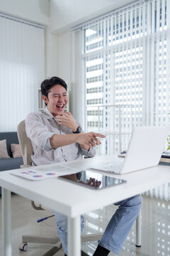 Young Asian businessman laughing during video call at home workspace pointing at laptop screen with copy space