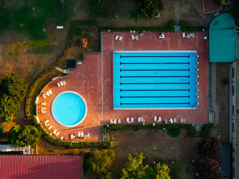 Aerial view of a swimming pool complex in the EUR district featuring a rectangular lap pool and a circular pool surrounded by lounge chairs and lush greenery in Rome, Lazio, Italy.