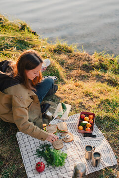 Woman preparing sandwiches and fresh vegetables for an outdoor picnic by a lake, arranging a simple lunch with reusable containers, mugs and thermos