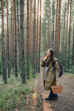 Woman examining a freshly picked wild mushroom while carrying a wicker basket on a pine forest trail during a seasonal mushroom hunting and foraging trip