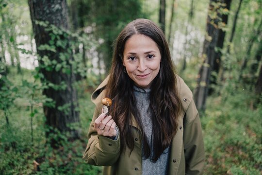 Woman holding a freshly picked wild mushroom and smiling at the camera in a green forest during a seasonal mushroom hunting and foraging trip in nature
