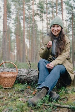 Smiling woman picking and tasting wild berries beside a wicker basket in a pine forest, enjoying seasonal foraging and a simple outdoor pleasure in nature