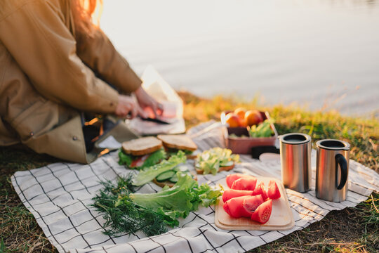 Woman preparing fresh picnic food outdoors by a lake, slicing ingredients for sandwiches with vegetables, fruit and reusable cups during a relaxed nature outing