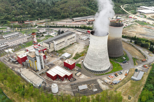 Aerial view of the Santa Barbara power plant with its iconic cooling towers emitting steam, surrounded by industrial buildings and lush green forest in Santa Barbara, Toscana, Italy.