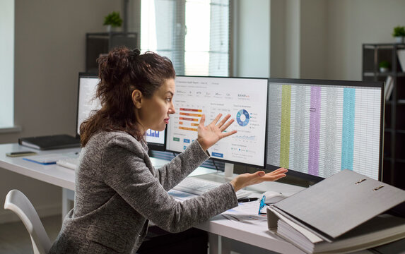 Woman analyst displays stress and frustration with data. She gestures at computer screens, questioning dashboard and charts, noting reporting errors and pressure. Clear concept of data crisis.