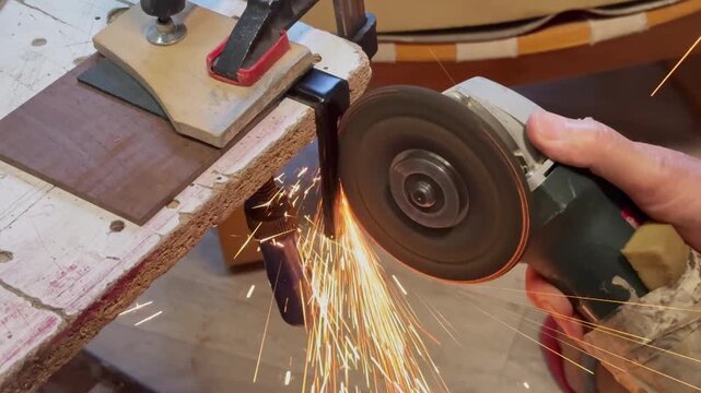 A side view of a craftsman using an electric power grinder to cut a steel bracket securely clamped to a workbench. Bright golden sparks illuminate the industrial workshop studio setting.