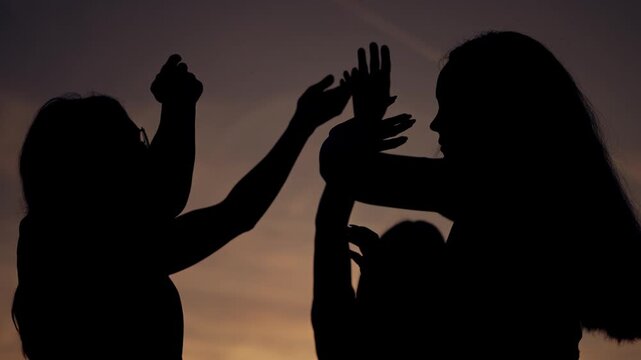 Two women silhouettes dance at sunset sky. Female friends raise hands in joyful movement against orange dusk. Girls enjoy outdoor freedom together. Sunset silhouettes show happy female bonding.