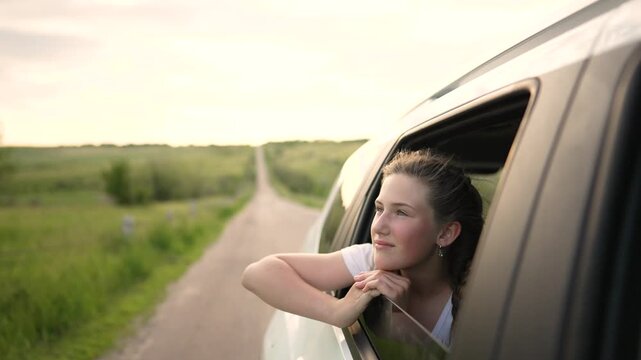 Young girl leaning out car window. Teenager enjoys road trip travel. Happy child looks at nature during summer vacation. Smiling girl rests on car window. Passenger on scenic country road drive.