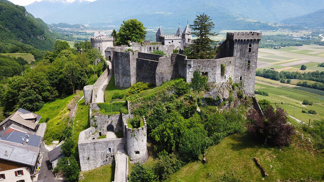 Aerial view of the historic Chateau de Miolans perched on a rocky cliff, surrounded by lush green trees and agricultural fields in Saint-Pierre-d'Albigny, Auvergne-Rhone-Alpes, France.