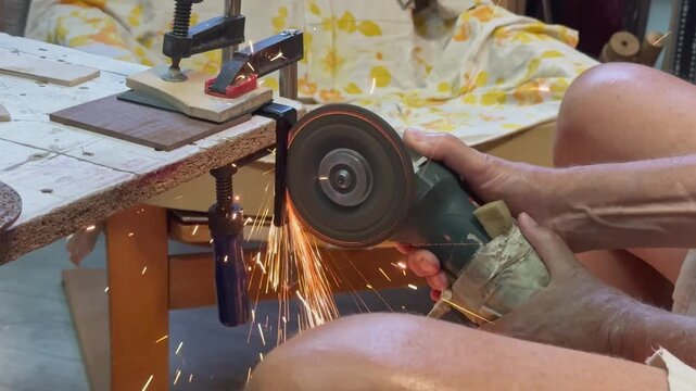 A side view of a craftsman using an electric power grinder to cut a steel bracket securely clamped to a workbench. Bright golden sparks illuminate the industrial workshop studio setting.