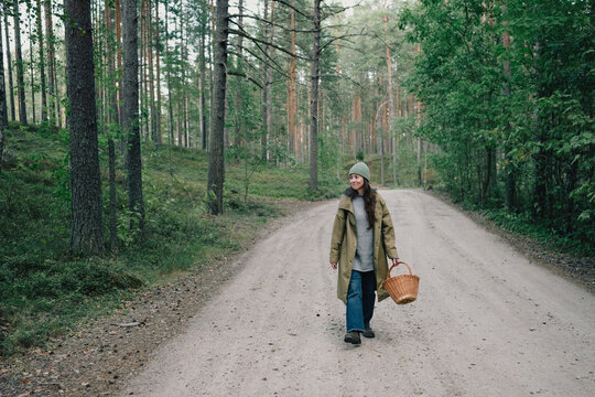 Woman carrying a wicker basket while walking along a quiet forest road, enjoying a slow outdoor lifestyle, solitude and peaceful time surrounded by nature