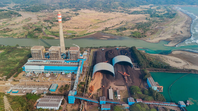 Aerial view of a large coal-fired power plant featuring a tall chimney, expansive coal storage domes, and industrial infrastructure situated along a river and the coastline