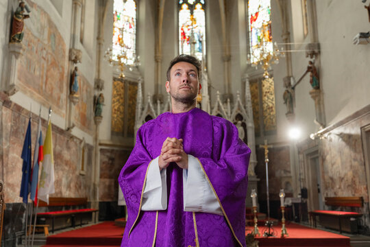 A catholic priest stands with hands clasped during a religious ceremony in a church in Europe. The setting features stained glass windows.