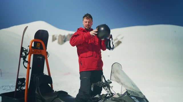A happy man removes his helmet, standing beside a snowmobile with a snowboard in snowy mountains. He smiles after a winter adventure on a bright, clear day.