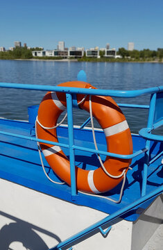 Bright orange lifebuoy ring hanging on a blue metal boat railing with a river and city skyline in the background.