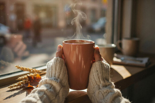 Person holding steaming ceramic mug wearing chunky knitted sweater by window