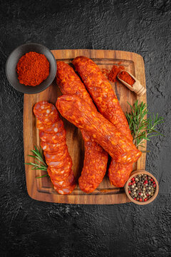 Sliced chorizo sausage arranged on a wooden cutting board with whole sausages, spices, and herbs on a textured black background