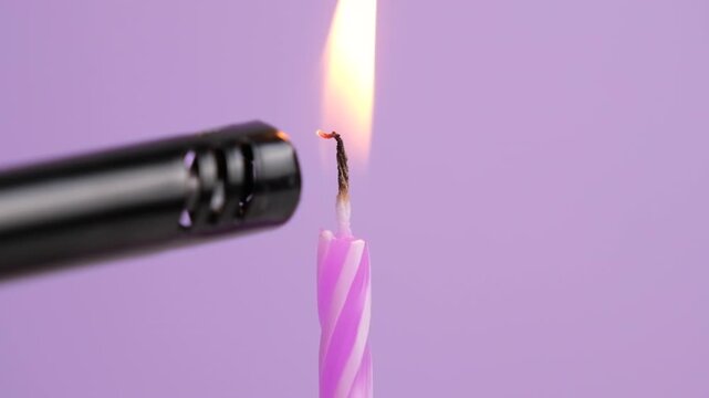 Pink striped birthday candle being lit with a lighter and burning with a bright flame against a soft purple background