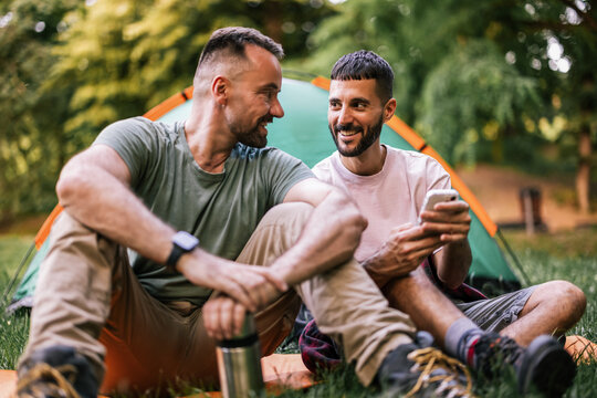 Gay couple sitting together and looking at a smartphone beside a camping tent