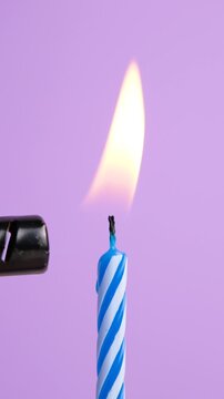 Blue striped birthday candle being lit with a lighter and burning with a bright flame against a soft purple background