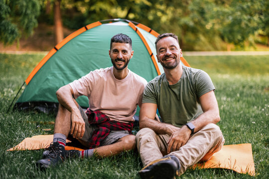 Gay couple sits together on the grass in front of their camping tent, smiling at the camera during an outdoor trip.
