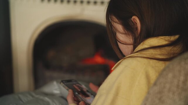 Focused girl watches social media video on phone while relaxing by fireplace