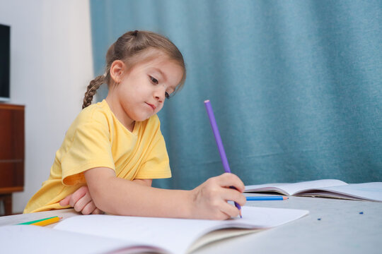 Young Girl With Braided Hair Concentrates While Drawing In Notebook At Home, Using Colored Pencils On Table Beside Open Sketchbook And Blue Curtain, Showing Quiet Indoor Learning