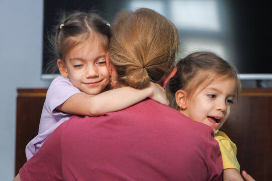 Young Mother Embraces Two Little Daughters In Close Family Hug Indoors, With Smiling Child Faces, Casual Home Clothing, Soft Natural Light, And Tender Everyday Comfort In Cozy Home