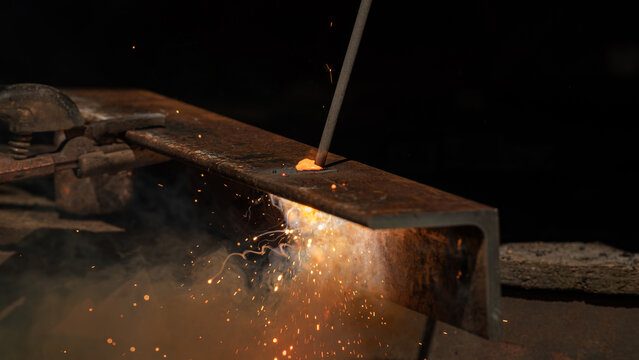 Close-up of arc welding on a steel structure with bright sparks, glowing metal and swirling smoke against a dark background. Industrial metalworking and fabrication process.
