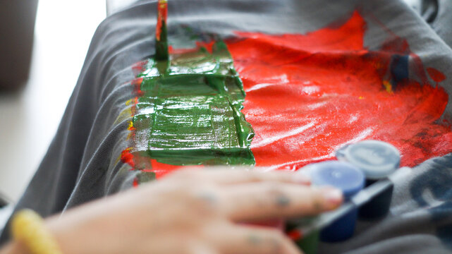 Child hands painting with acrylic colors on a grey t-shirt at home workspace