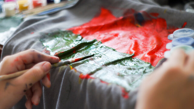 Child hands painting with acrylic colors on a grey t-shirt at home workspace