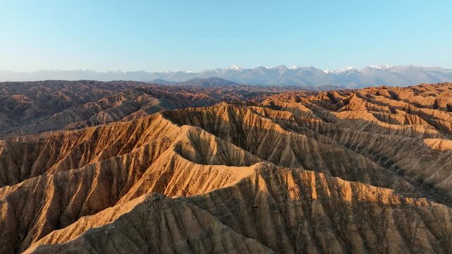 Aerial view of the rugged Ak Say canyon landscape with dramatic golden hour lighting and distant snow-capped mountains in Ak Say, Issyk-Kul Region, Kyrgyzstan.