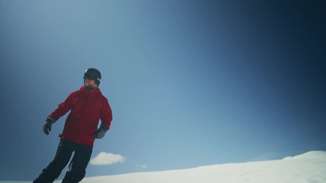 A person in a red jacket snowboards down a wide, snow-covered mountain slope. Another person on a snowboard follows, enjoying the sunny winter day under a clear blue sky.