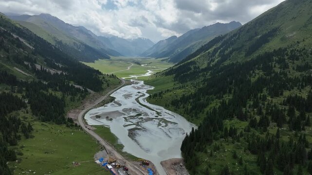 Aerial view of Chon Ashu Pass featuring a winding river through a lush mountain valley with pine forests and dramatic peaks in Karakol, Issyk-Kul Region, Kyrgyzstan.