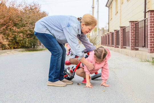 Mature Woman Bends Down To Comfort Small Girl Who Has Fallen From Red Bicycle, Offering Steady Support On Neighborhood Street Near Houses And Autumn Trees, Showing Family Care