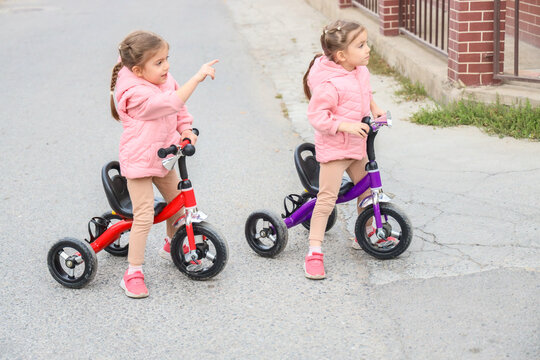 Two Little Girls Ride Colorful Tricycles Beside Brick Houses On Quiet Neighborhood Street, One Pointing Forward While Both Wear Pink Jackets And Casual Shoes