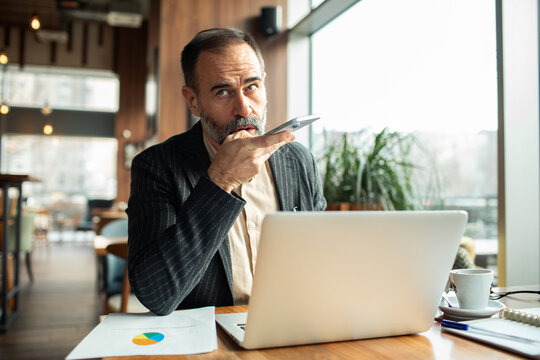 Mature businessman using voice assistant on smartphone at cafe workspace