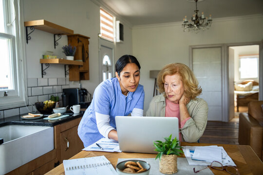 Caregiver helping senior woman use laptop in home kitchen