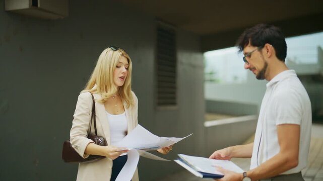 Two business people engage in a heated discussion outdoors, expressing frustration over documents. The man and woman display clear disagreement during their professional encounter.