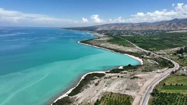 Aerial view of the turquoise waters of Issyk Kul lake, with a coastal road winding through green fields and mountains in Cholpon Ata, Issyk-Kul Region, Kyrgyzstan.